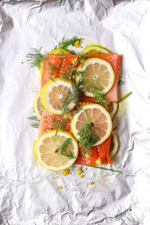 Fresh salmon fillet topped with lemon slices, dill, and mustard seeds on crinkled parchment paper, ready for cooking or serving.