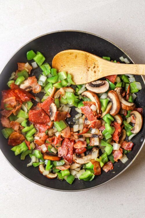 Sautéed mushrooms, bacon, celery, and onions in a black skillet with a wooden spoon.