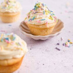 Colorful sprinkle-topped cupcakes on a white plate in a light kitchen setting.