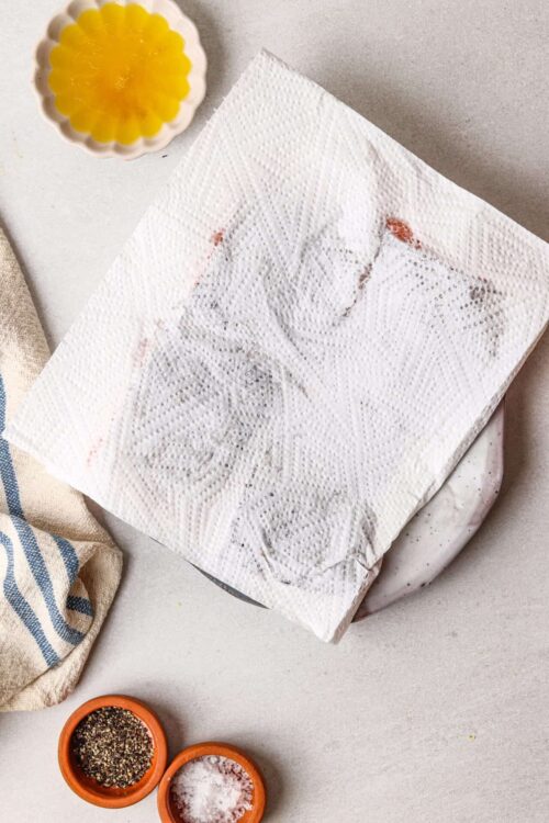 Seasoned raw chicken breast with paper towel covering, on a baking tray for food preparation.