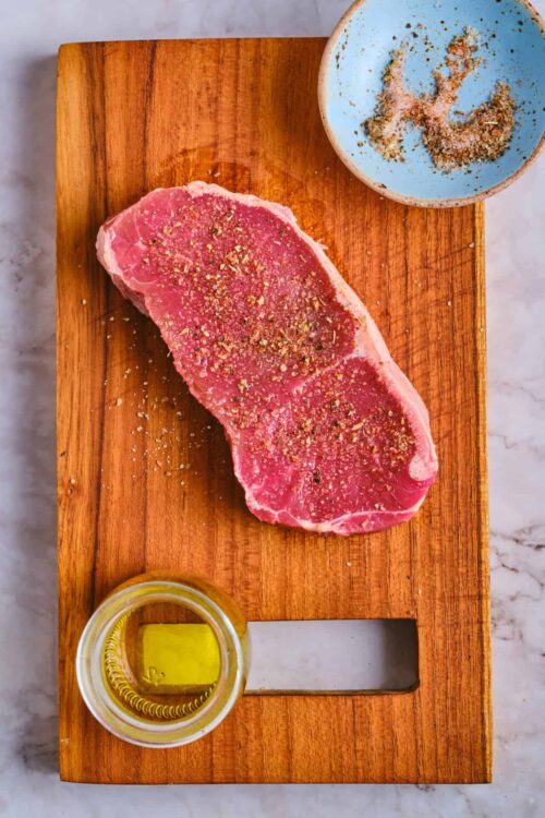 Juicy raw steak on wooden cutting board with sea salt and herbs, olive oil and seasoning bowl, high-quality meat preparation for cooking or grilling.