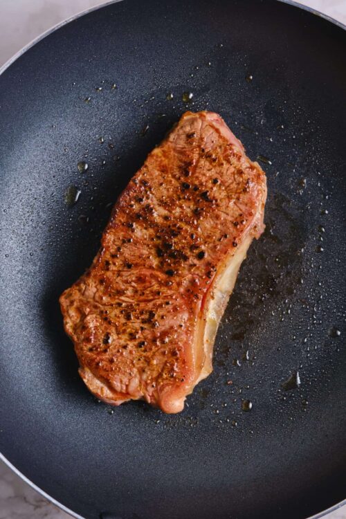 Juicy pan-seared steak cooking in a non-stick skillet with seasoned crust and sizzling oil.