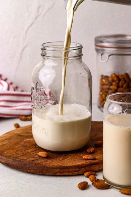 Creamy almond milk being poured into a jar on a wooden board, with almonds in the background.