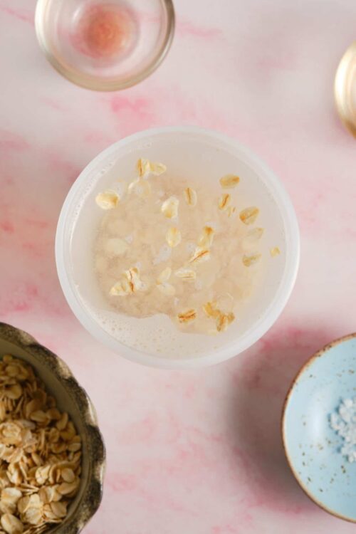 Cream of oats in a glass bowl with oats and ingredients around it, on a pink background.