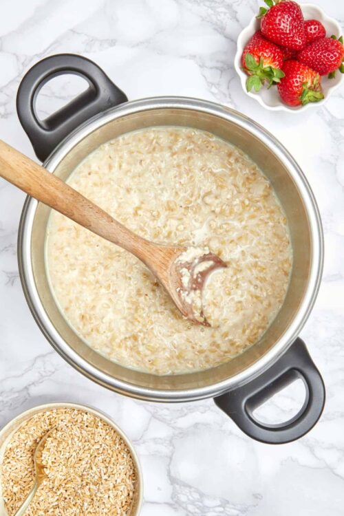 Creamy oatmeal cooking in a pot with a wooden spoon, with strawberries in a bowl nearby.
