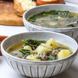 Hearty potato and sausage soup in textured white bowls with fresh ingredients in the background.
