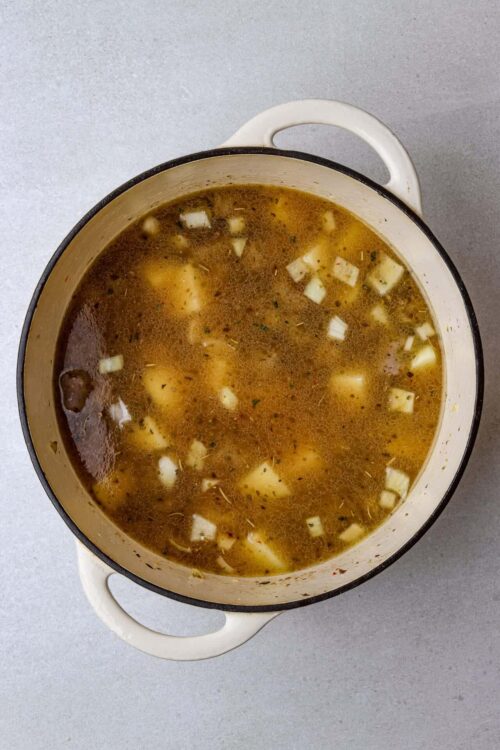 Hearty beef and vegetable soup in a white enamel pot with black rim on a light gray surface.