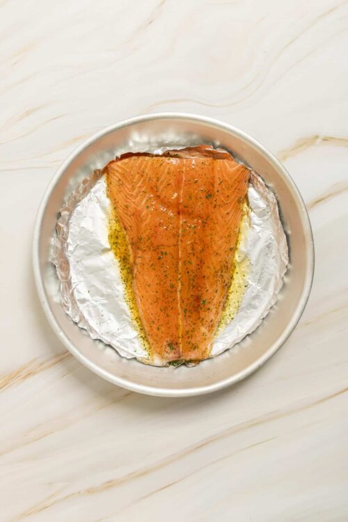 Fresh salmon fillet seasoned with herbs on aluminum foil in a baking dish.