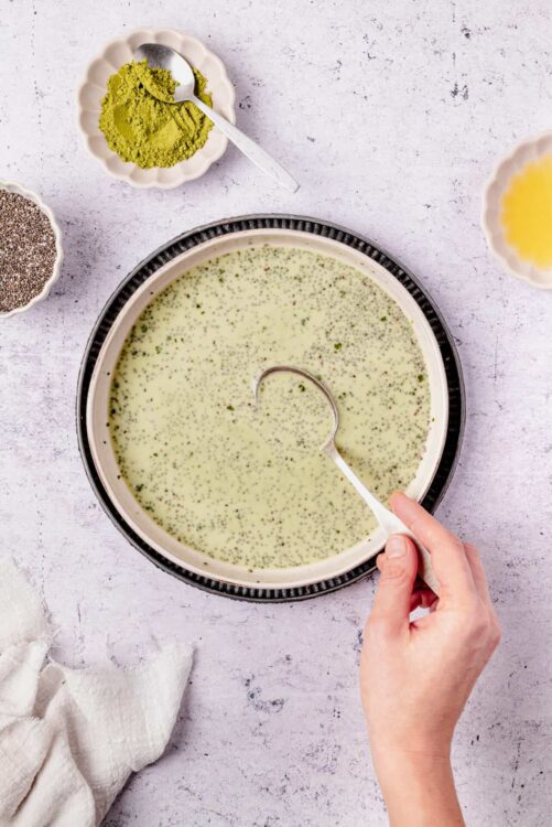 Matcha green tea soup in a bowl with a spoon, surrounded by matcha powder, chia seeds, and lemon zest.