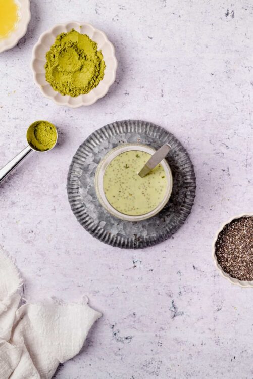 Bright green matcha powder in a scalloped dish and spoon with a creamy matcha latte on a textured gray plate surrounded by ingredients.