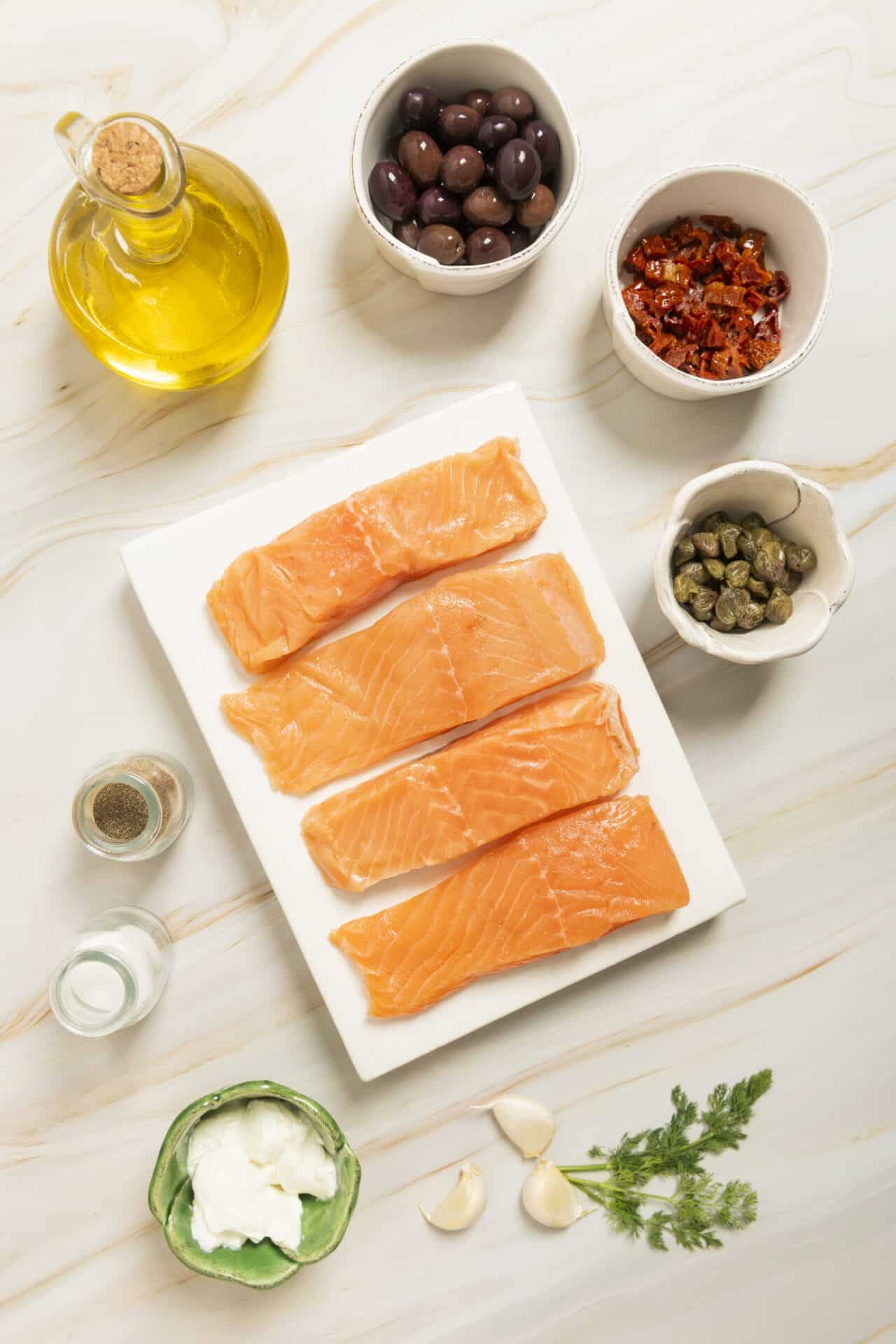 Fresh salmon fillets with capers, sun-dried tomatoes, black olives, and garlic on a marble countertop for healthy seafood recipes.