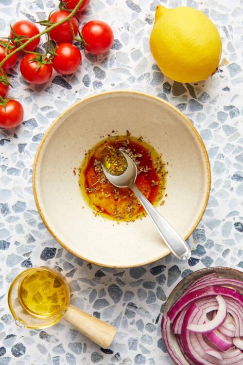 Fresh cherry tomatoes on vine, whole lemon, sliced red onion, and olive oil with herbs on a mosaic surface.