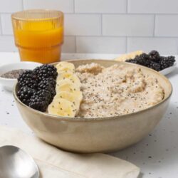 Creamy oatmeal in a beige bowl topped with banana slices and blackberries, served with fresh fruit and chia seeds.