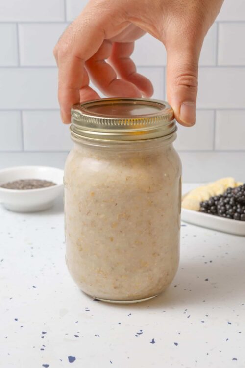 Homemade oatmeal in a mason jar on a white countertop with baking ingredients in the background.