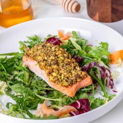Roasted salmon with chopped pistachio crust on mixed greens salad in white bowl.