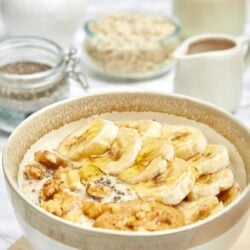 Creamy banana pudding in a white bowl with walnuts and vanilla wafers, surrounded by ingredients on a light wooden surface.