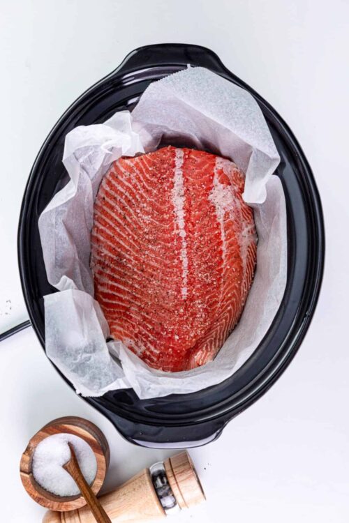 Juice salmon fillet seasoned with salt and spices in a black slow cooker with parchment paper.