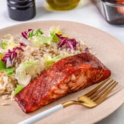 Salmon with rice and salad on a beige plate, olive oil, soy sauce, and baked dish in background.