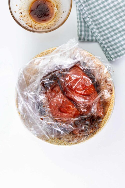 Marinated tomatoes wrapped in plastic on a patterned plate with balsamic vinegar in a glass bowl and a green checkered napkin.