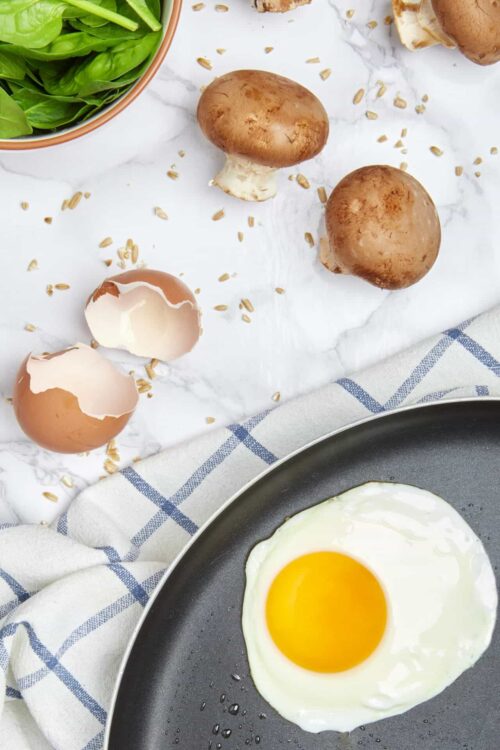 Fresh mushrooms, cracked eggs, and spinach on white marble surface.