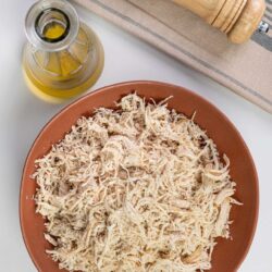 Shredded cooked chicken in a terracotta bowl with olive oil, salt grinder, and striped cloth on white surface.
