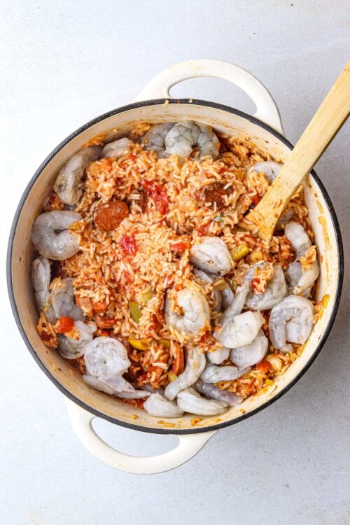 Shrimp paella in a white skillet with a wooden spoon, featuring rice, vegetables, and seafood.