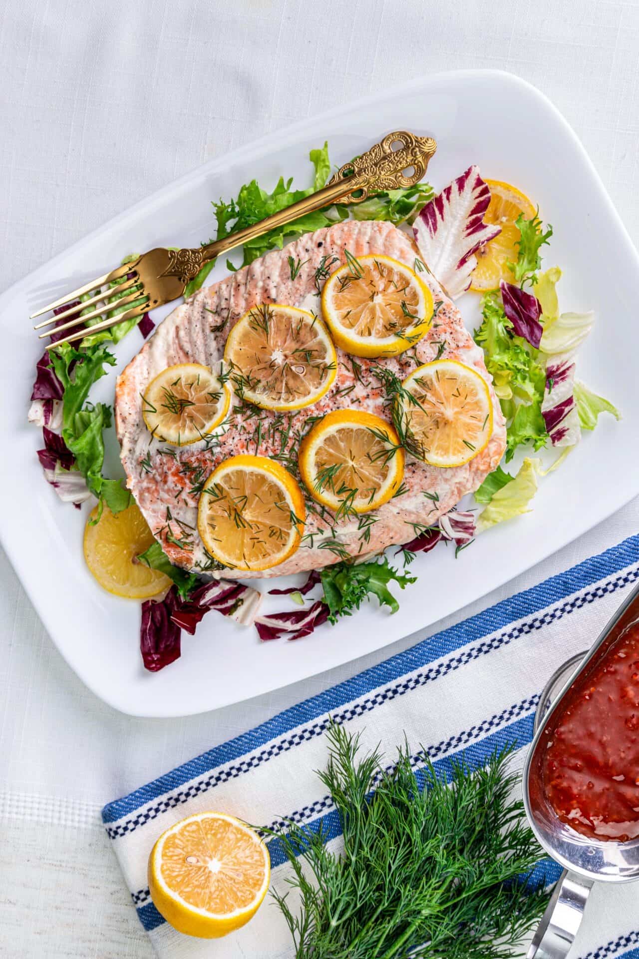 Fresh lemon herb baked fish on a white platter with leafy greens, garnished with lemon slices and dill, served on a white table with a blue and white striped cloth.