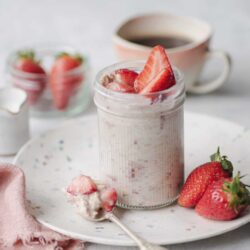 Fresh strawberry smoothie in a glass jar with sliced strawberries on top, served on a white plate with whole strawberries and a pink napkin.