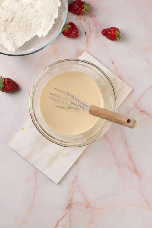 Creamy vanilla custard mixture in glass bowl with whisk and fresh strawberries on marble surface.