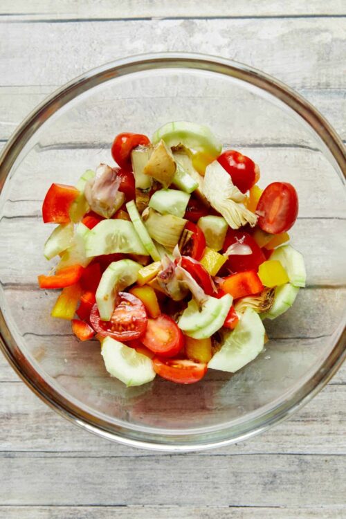 Fresh vegetable salad with cherry tomatoes, cucumbers, bell peppers, and artichokes in glass bowl.
