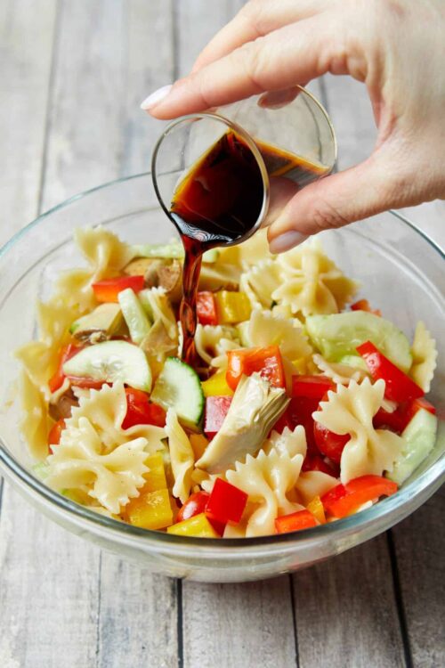 Golden bolognese sauce being poured over vegetable pasta salad in a glass bowl.