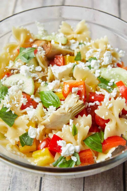 Colorful pasta salad with vegetables, feta cheese, and fresh basil in a glass bowl.
