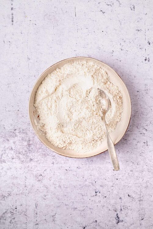 Flour in a beige bowl on textured white surface, ideal for baking and recipe preparation.