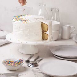 Delicious white frosted birthday cake with sprinkles on top, being decorated on a white cake stand.
