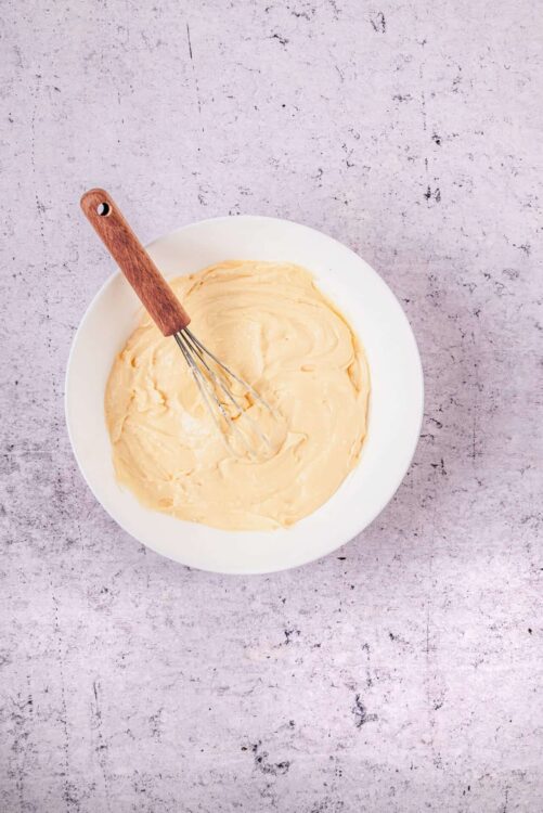 Creamy homemade pastry dough in a white bowl with a whisk on a rustic surface.