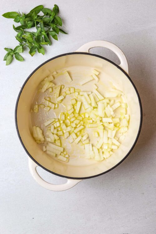 Sautéed diced onions in a cream-colored cast iron skillet with fresh basil on a white surface, cooking preparation for baked good recipes.