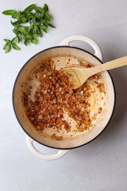 Sautéed onion and herb mixture in a creamy enameled cast iron pot with a wooden spoon and fresh basil.