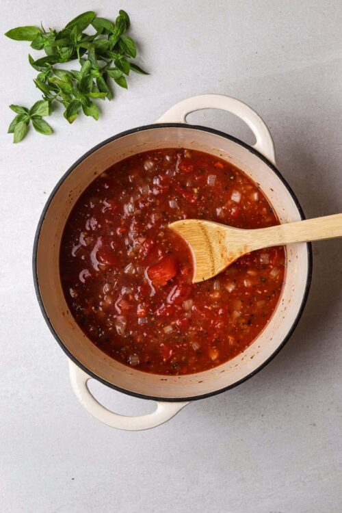 Savory homemade tomato soup in a white enamel pot with a wooden spoon and fresh basil.