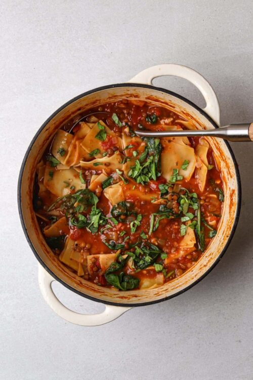 Hearty vegetable lasagna stew in a white enameled cast iron pot, garnished with fresh herbs. Perfect comfort food recipe from Baked Bree.