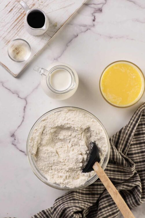 Flour, milk, and vanilla extract for baking on white marble surface.