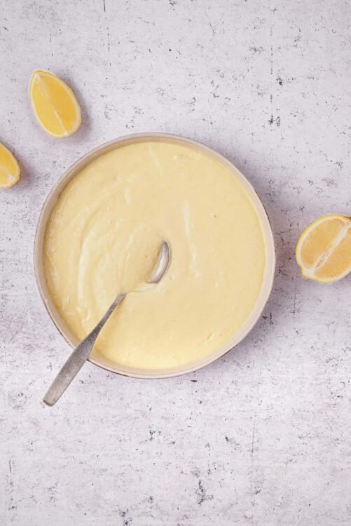 Lemon curd in a bowl with a spoon and lemon wedges on a gray textured surface.