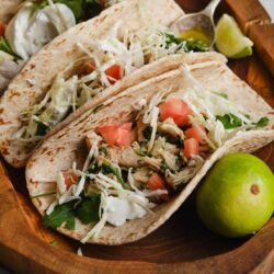 Fresh chicken tacos with shredded cabbage, tomatoes, and cilantro on flour tortillas, served on a wooden tray with lime wedges.