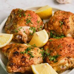 Crispy roasted chicken thighs with lemon wedges and fresh herbs on white ceramic plate.