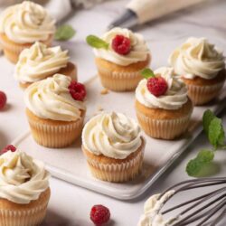 Butter icing cupcakes topped with raspberries and mint leaves on a white platter.
