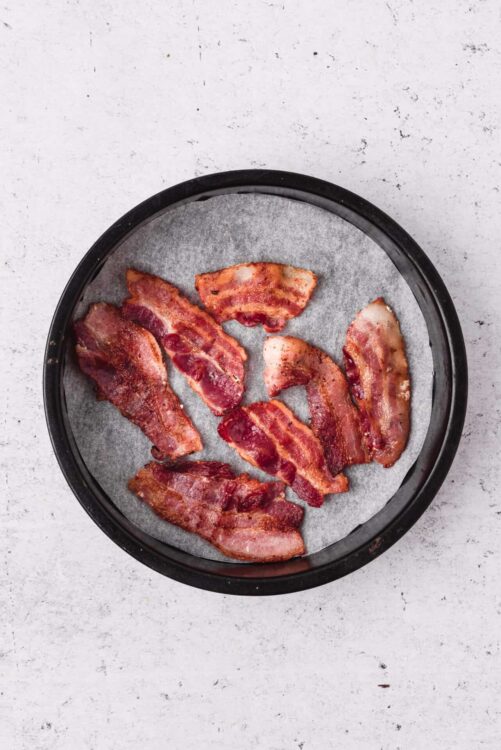 Crispy baked bacon slices on parchment paper in a black round baking tray.