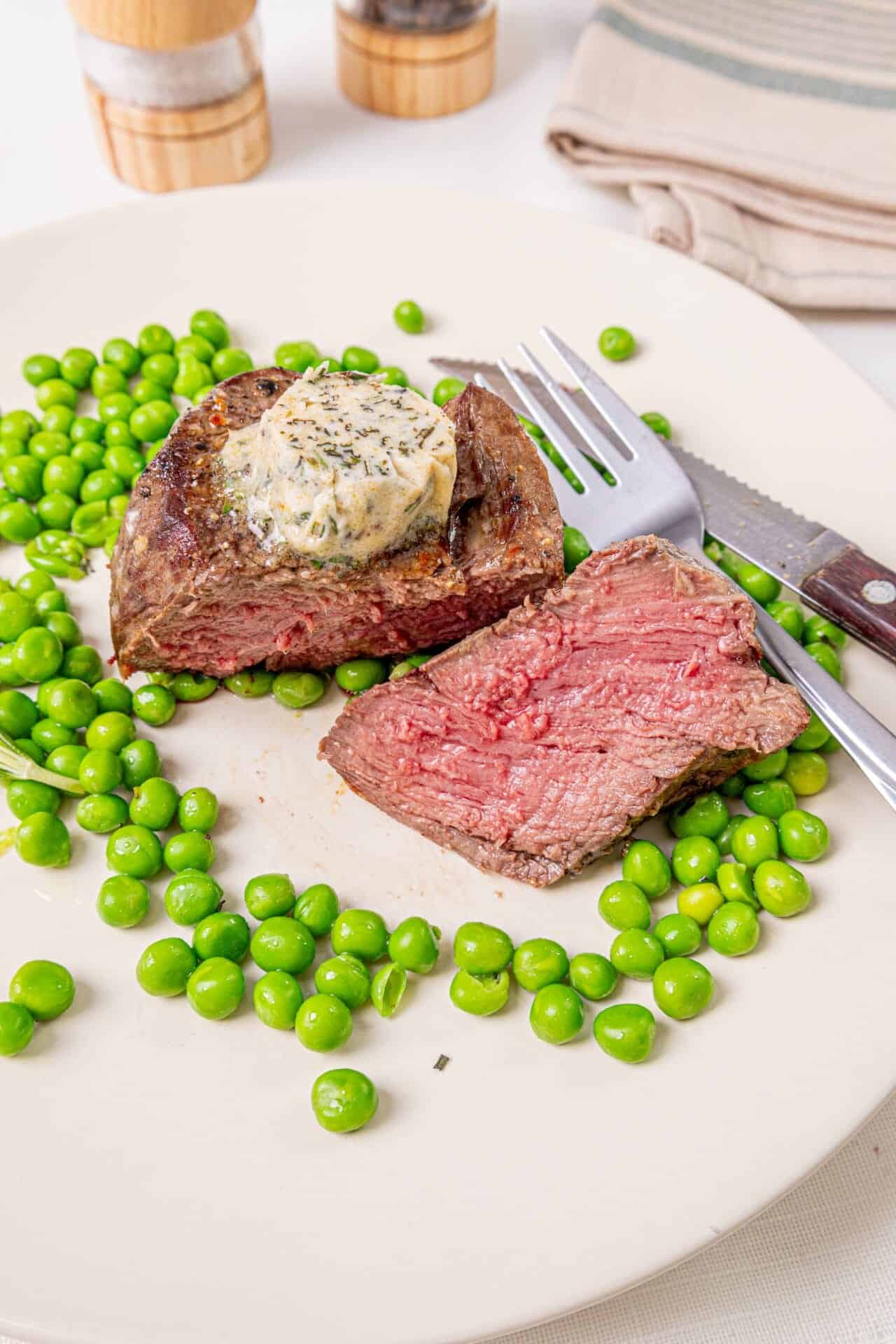 Juicy beef filet mignon with herb butter, served with fresh green peas on a white plate.