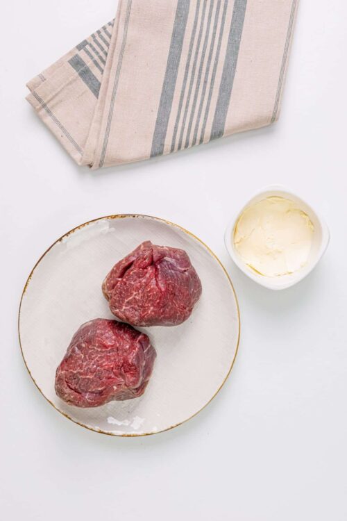 Juicy raw beef steaks with butter on a white plate, kitchen towel in background, fresh ingredients for cooking.