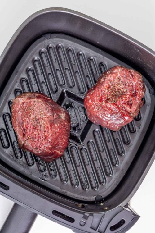 Raw steak seasoned with salt and pepper in an air fryer basket.