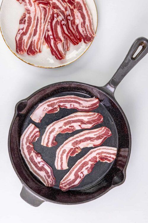 Bacon strips cooking in a skillet with raw bacon slices on a plate above.