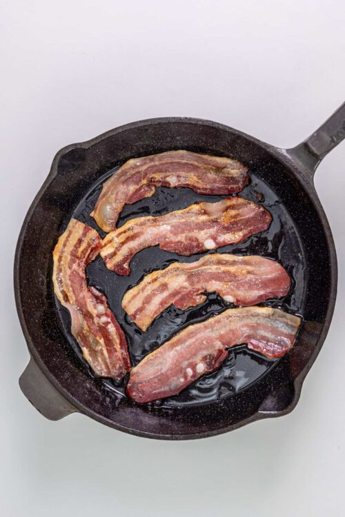Crispy bacon frying in a black skillet on a white background.